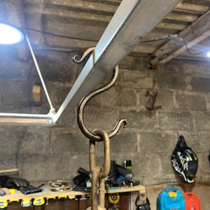 May include: A close-up of a heavy-duty metal hook attached to a metal beam. The hook is connected to a chain, which is hanging down. The hook is designed for hanging heavy objects.