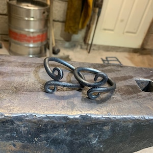 May include: Two handcrafted, dark metal rings with a swirling design, resting on a weathered anvil. The rings have a rustic, forged appearance, showcasing the artistry of metalwork. The background suggests a workshop setting.