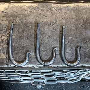 May include: Three metal hooks, each with a unique shape, are displayed on a textured surface. The hooks are made of a dark metal and have a rustic appearance.