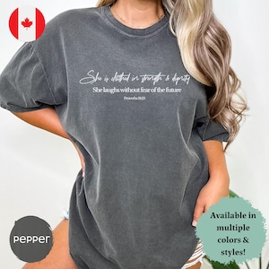 Peut inclure: Un t-shirt surdimensionné gris foncé avec un imprimé graphique blanc qui dit "She is clothed in strength & dignity She laughs without fear of the future Proverbs 31:25".