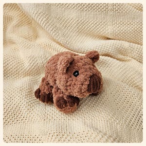 May include: A brown plush toy shaped like a pig with a textured surface. The toy is lying on a cream-colored knitted blanket.