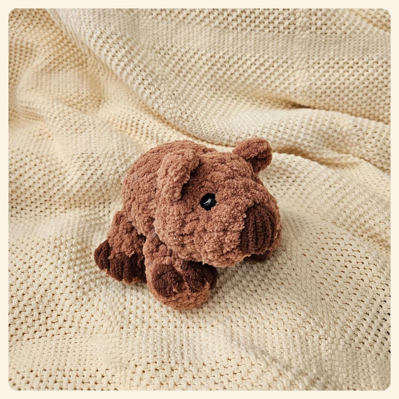 May include: A brown plush toy shaped like a pig with a textured surface. The toy is lying on a cream-colored knitted blanket.