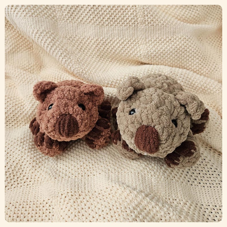 May include: Two plush brown and beige wombat toys on a cream knitted blanket. The wombats have a textured, fuzzy surface and are lying on their bellies.