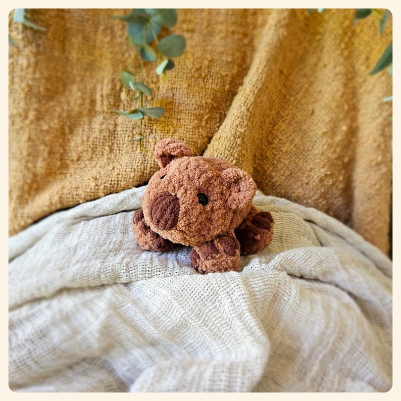 May include: A brown crocheted plush toy shaped like a wombat, lying on a white blanket.