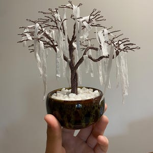 May include: A wire sculpture of a tree with white fabric hanging from the branches. The tree is in a brown and green ceramic pot with white stones.
