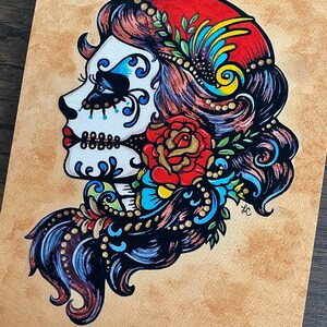 May include: A colorful illustration of a Day of the Dead sugar skull with a red rose, blue and gold floral accents, and a red and yellow headdress.