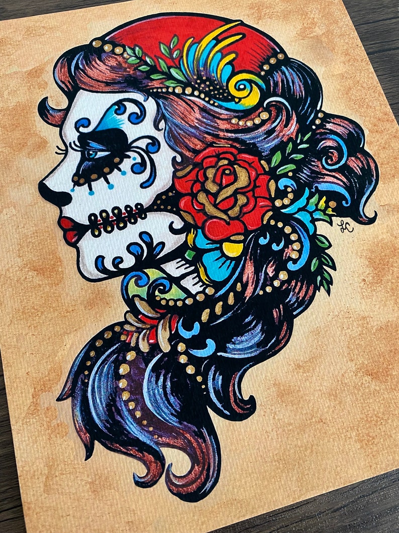 May include: A colorful illustration of a Day of the Dead sugar skull woman with a red rose and floral details. The skull is painted in white with blue and black accents. The woman's hair is black with blue and gold highlights. The background is a light brown.