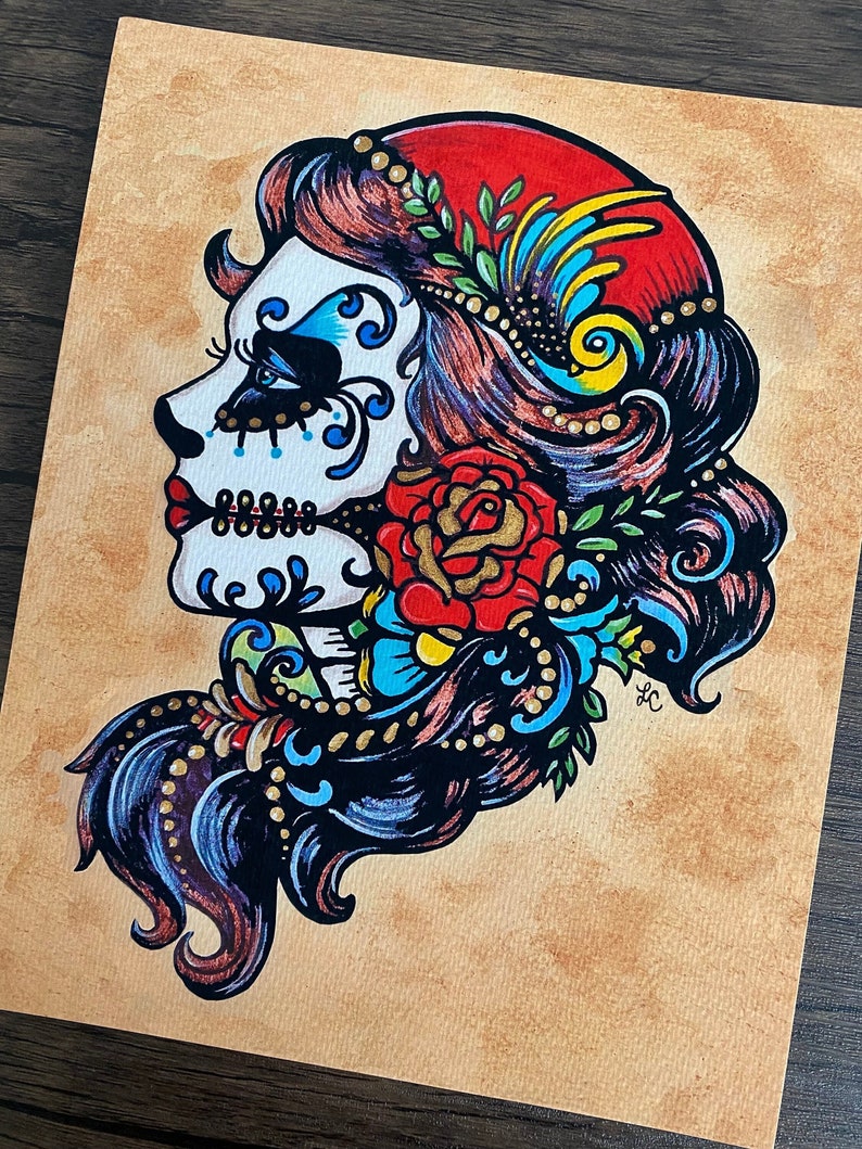 May include: A colorful illustration of a Day of the Dead sugar skull with a red rose, blue and gold floral accents, and a red and yellow headdress.