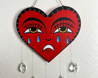 Peut inclure: Décoration murale en forme de cœur rouge avec un visage triste. Le cœur présente des sourcils noirs, des cils et une bordure noire avec des accents turquoise. Des larmes de cristal transparentes pendent du bas, suspendues par de fins fils. Une chaîne noire est utilisée pour l'accrochage.