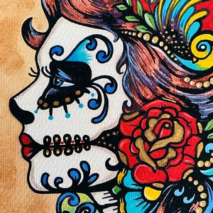 May include: A colorful illustration of a woman with a sugar skull face, red lips, and a red rose. The woman is wearing a red head scarf with gold beads and a blue and yellow feather. The background is a light brown.