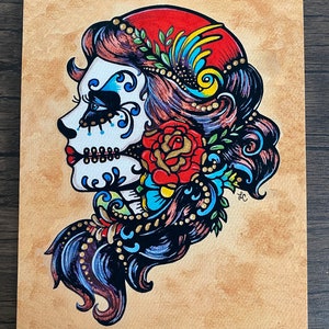 May include: A colorful illustration of a Day of the Dead sugar skull woman with a red flower and blue and gold floral details. The skull is white with black and blue details. The woman has long dark hair with red and blue highlights.