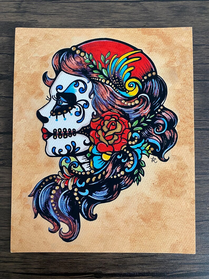May include: A colorful illustration of a Day of the Dead sugar skull woman with a red flower and blue and gold floral details. The skull is white with black and blue details. The woman has long dark hair with red and blue highlights.