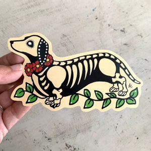 Puede incluir: Una ilustración en blanco y negro de un esqueleto de dachshund con una guirnalda de flores rojas. El dachshund camina sobre una vid verde con hojas.