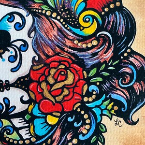 May include: A colorful illustration of a sugar skull with a red rose, blue and green floral details, and a yellow bird. The skull is surrounded by black hair with gold beads.