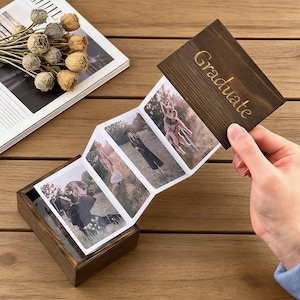 May include: A dark brown wooden photo box with a hinged lid, displaying a series of graduation photos. The lid is engraved with the word "Graduate" in gold lettering. The photos are in a concertina style, and the box is open, revealing more photos.