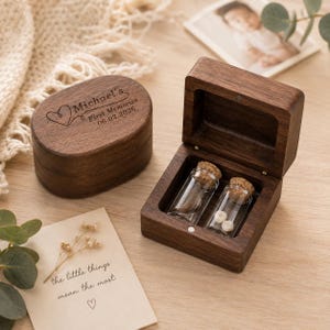 May include: A dark wood keepsake box with a hinged lid, engraved with the text Michael's First Memories 06.02.2026 and a heart icon. Inside the open box are two small glass vials with cork stoppers, one containing two small white teeth.