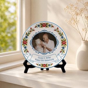 Peut inclure: Une assiette décorative ronde blanche présentant une photographie centrale en noir et blanc d'un couple âgé. L'assiette est ornée d'une bordure florale colorée de roses rouges et de fleurs bleues, et d'une bannière avec le texte Eleanor & Arthur Happy 50th Anniversary.