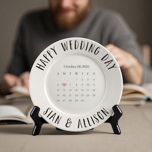 Op de afbeelding: Een witte keramische plaat met de tekst "HAPPY WEDDING DAY" en een kalender voor oktober 2025. De trouwdatum is gemarkeerd met een hart. De namen "SIAN & ALLISON" staan eronder. De plaat staat op een zwarte standaard.