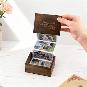 以下が含まれることがあります： ダークブラウンの木製フォトボックス。ヒンジ付きの蓋には、一連の写真が取り付けられています。蓋には「Jason & Eli」の名前、ハート、そして「Our First Year 12.10.2025」の文字が刻まれています。ボックスは開いており、写真が表示されています。
