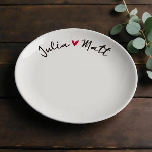 Może przedstawiać: Owalny biały ceramiczny talerz z imionami "Julia" i "Matt" w czarnym skrypcie, oddzielonymi czerwonym sercem. Talerz leży na ciemnej drewnianej powierzchni. Gałązki eukaliptusa w tle.