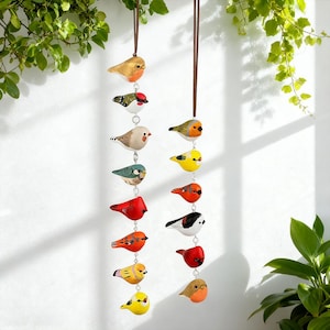 May include: A decorative hanging of painted bird ornaments in various colours, including red, orange, yellow, and blue. The birds are suspended from brown cords against a white background, with green foliage in the corners. Each bird has a unique design.