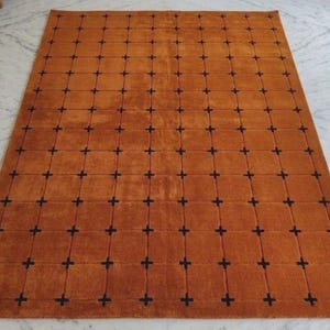 Puede incluir: Una alfombra rectangular con un fondo de color naranja quemado sólido y un patrón de cuadrícula repetitivo. La cuadrícula está formada por líneas finas verticales y horizontales de color marrón oscuro con pequeñas cruces.