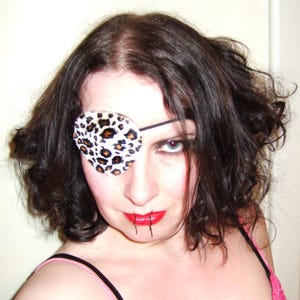 May include: A woman with dark brown hair is wearing a leopard print eye patch over her right eye. She is wearing a pink and white tank top and has red lipstick on.