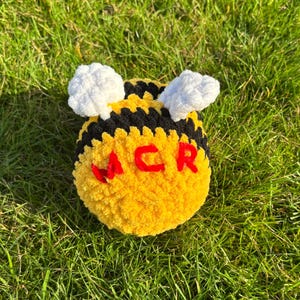 Könnte beinhalten: Ein rundes, gehäkeltes Bienen-Plüschtier mit gelben und schwarzen horizontalen Streifen, zwei weißen, strukturierten Flügeln auf der Oberseite und den roten, gestickten Buchstaben MCR auf der Vorderseite aus weichem Garn.