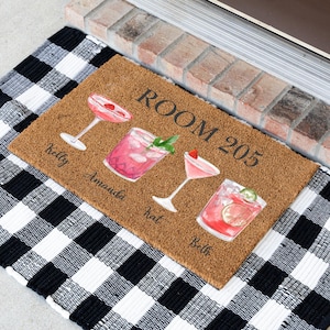 May include: A rectangular doormat with a brown coir surface features four watercolor cocktail illustrations and the text "ROOM 205". Names "Kelly", "Amanda", "Kat", and "Beth" are below the cocktails. The mat rests on a black and white checkered rug.
