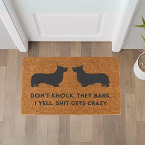 May include: A rectangular doormat with a brown background and two black corgi silhouettes facing each other. The text "DON'T KNOCK. THEY BARK. I YELL. SHIT GETS CRAZY" is printed in black.