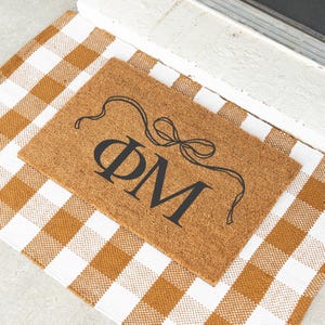 May include: A rectangular coir doormat with the letters ΦM in black, topped with a decorative bow design. The doormat is placed on a brown and white checkered rug, creating a welcoming entrance.