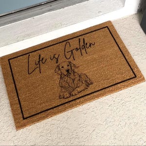 May include: A brown coir doormat with a black border and the text "Life is Golden" in black script. A black and white illustration of a golden retriever dog is centered on the mat.