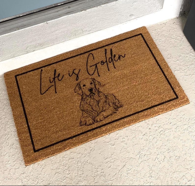 May include: A brown coir doormat with a black border and the text "Life is Golden" in black script. A black and white illustration of a golden retriever dog is centered on the mat.