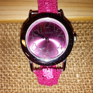 May include: A pink wristwatch with a silver face and black hands. The watch face has the word "NARMI" printed on it. The watch band is made of pink braided fabric.
