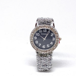 Puede incluir: Un reloj plateado con una esfera negra y agujas plateadas. El reloj tiene una correa plateada con un diseño trenzado y diamantes de imitación alrededor de la esfera. La esfera del reloj tiene la palabra "NARM" impresa en ella.