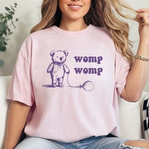 Puede incluir: Una camiseta rosa de gran tamaño con un oso de dibujos animados que sostiene un globo. La camiseta tiene el texto "womp womp" impreso en ella.
