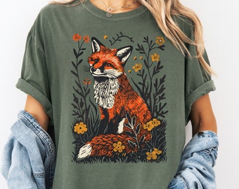 以下が含まれることがあります： 花畑に座っているキツネのグラフィックが描かれた緑色のTシャツ。キツネはオレンジ色と白で、目は黒。花は黄色とオレンジ色です。