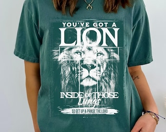 May include: A teal green t-shirt with a white graphic print of a lion's face with the text "You've got a lion inside of those lungs. So get up & praise the Lord."