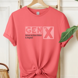 May include: Coral-colored t-shirt with the text "GEN X" in a distressed, light pink font. Below the text, it reads "Raised on Hose Water & Neglect." The shirt is knotted at the waist and hangs on a wooden hanger.