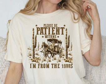 Puede incluir: Una camiseta beige con un gráfico marrón de un carruaje tirado por caballos en un paisaje desértico. El texto "Please be patient with me I'm from the 1900s" está impreso en la camiseta.