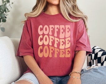 Op de afbeelding: Een vrouw draagt een rood T-shirt met het woord "COFFEE" dat drie keer wordt herhaald in een retro-lettertype. Het shirt is roodvervaagd en de tekst is in tinten roze en oranje.