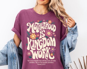 May include: A purple t-shirt with a graphic design that says "Motherhood is Kingdom Work" in a retro font with flowers surrounding the text. The quote "She is strong and dignified, laughs at the future. Speaks with wisdom and kindness. Takes care of her household, avoids idleness. Proverbs 31:25-27" is printed below the graphic.