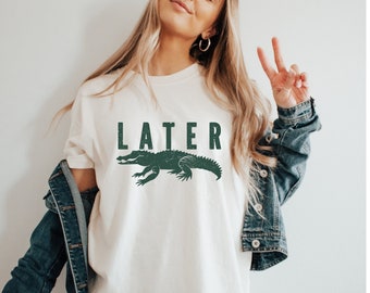 Peut inclure: T-shirt blanc avec un graphique d'alligator vert et le mot "LATER" imprimé en noir.