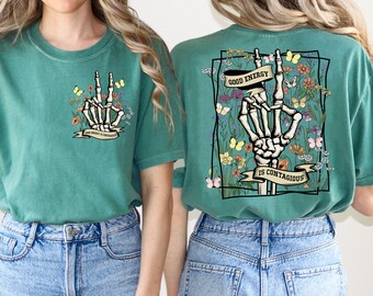 May include: A teal green t-shirt with a skeleton hand making a peace sign surrounded by flowers and the text "Good energy is contagious".
