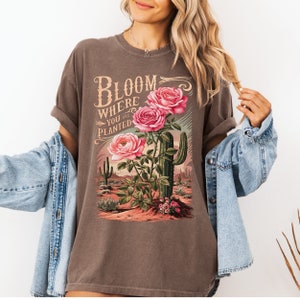 May include: A brown t-shirt with a graphic print of pink roses and a cactus. The text "Bloom Where You Are Planted" is printed on the shirt.