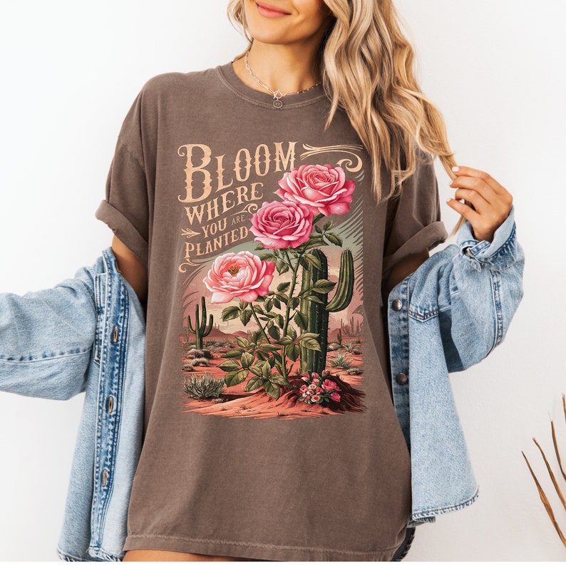 May include: A brown t-shirt with a graphic print of pink roses and a cactus. The text "Bloom Where You Are Planted" is printed on the shirt.