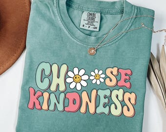 May include: A teal blue t-shirt with the words "Choose Kindness" in colorful, retro-style lettering. Two white daisies with yellow smiley faces are between the words "Choose" and "Kindness".