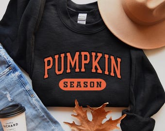May include: Black sweatshirt with the text "Pumpkin Season" in orange letters. The sweatshirt is laid out on a surface with a brown hat and a fall leaf.