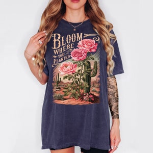 May include: A dark blue t-shirt with a vintage-style graphic of pink roses and a cactus. The graphic says "Bloom Where You Are Planted."
