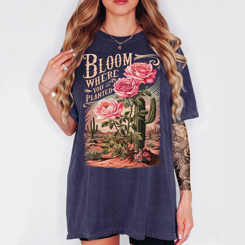 May include: A dark blue t-shirt with a vintage-style graphic of pink roses and a cactus. The graphic says "Bloom Where You Are Planted."